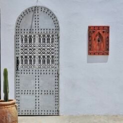 Red vintage Moroccan wooden panel hung on white wall next to traditional arched metal door with cactus pot